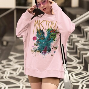 May include: Light pink sweatshirt with the word "MASTODON" in gold lettering. The front features a colorful graphic design of a creature with multiple eyes and tentacles. The person is wearing pink sunglasses.