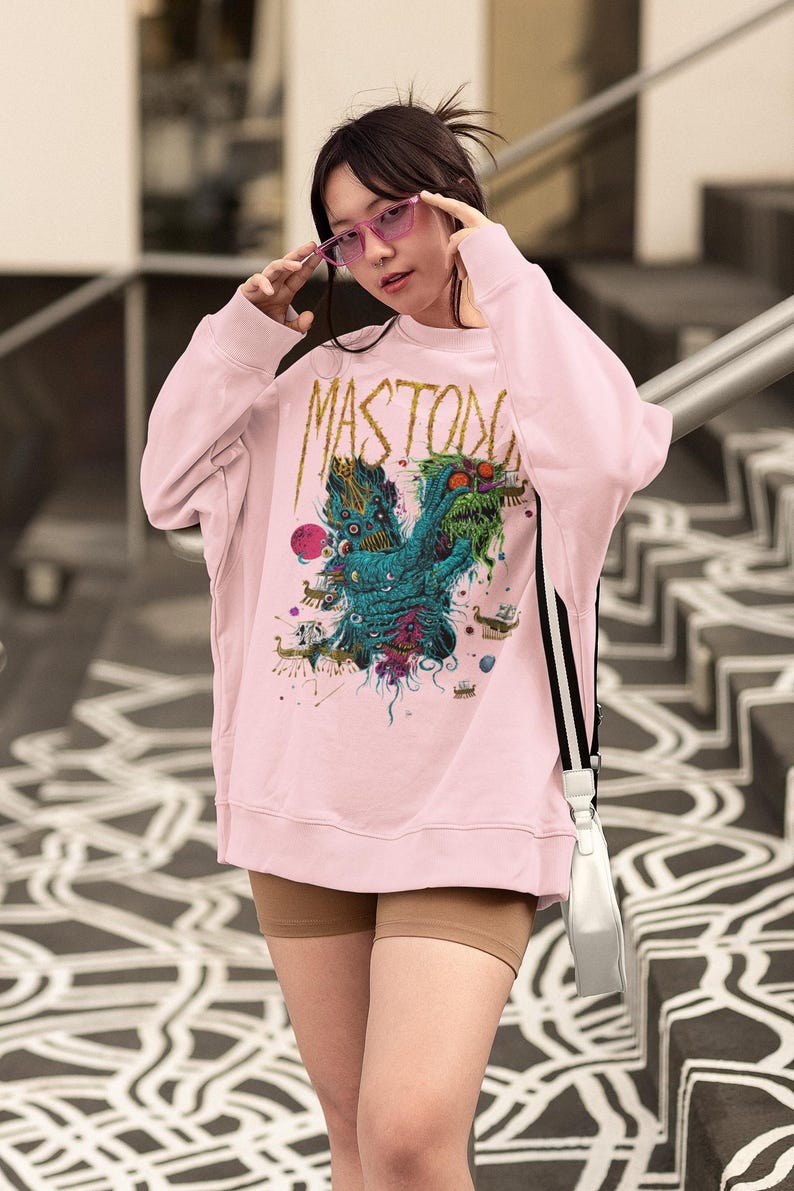 May include: Light pink sweatshirt with the word "MASTODON" in gold lettering. The front features a colorful graphic design of a creature with multiple eyes and tentacles. The person is wearing pink sunglasses.