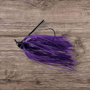 May include: A purple fishing jig with a black hook and head. The jig features a skirt of long, thin, purple strands, and is designed for attracting fish. The background is a wooden surface.