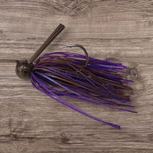 May include: A fishing jig with a dark brown head, a large hook, and a skirt of purple and brown strands. The jig has a bundle of brown bristles extending from the head.