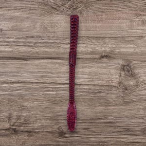 May include: A soft plastic fishing lure in a deep red color with a textured body and a slender, ribbed tail. The lure has a speckled appearance and is designed to mimic a worm or other aquatic prey.