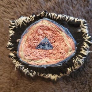 May include: A close-up of a handmade embroidered patch with a pink, white, and blue triangle design. The patch is surrounded by a black fabric border with white stitching.