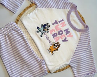May include: A long-sleeve shirt and pants set featuring white and light purple horizontal stripes. The shirt has a white center panel with embroidered cartoon characters and the text "IN MY TWOEY ERA" and "Lottie-Mae" in purple and pink.