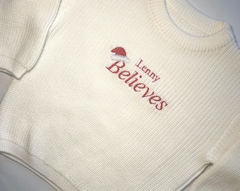May include: Cream-colored knit sweater with the embroidered text "Lenny Believes" in red, accompanied by a Santa hat graphic. The sweater features a ribbed crew neck and cuffs.