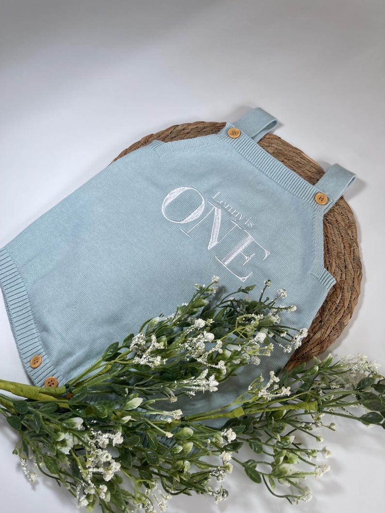 May include: A light blue knitted romper with the text "Lenny's ONE" in white. The romper has wooden buttons on the straps and sides. The romper is displayed on a woven mat with a sprig of white flowers.