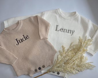 May include: Two knitted baby outfits, one beige and one white, each with a name embroidered on the front. The beige outfit is a romper with buttons. The white outfit is a sweater. A sprig of dried ornamental grass is in the foreground.