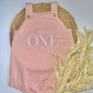 May include: A light pink knitted romper with the embroidered text "Daisy's ONE". The romper has wooden buttons on the straps and at the bottom. The romper is displayed on a woven mat with dried ornamental grass.