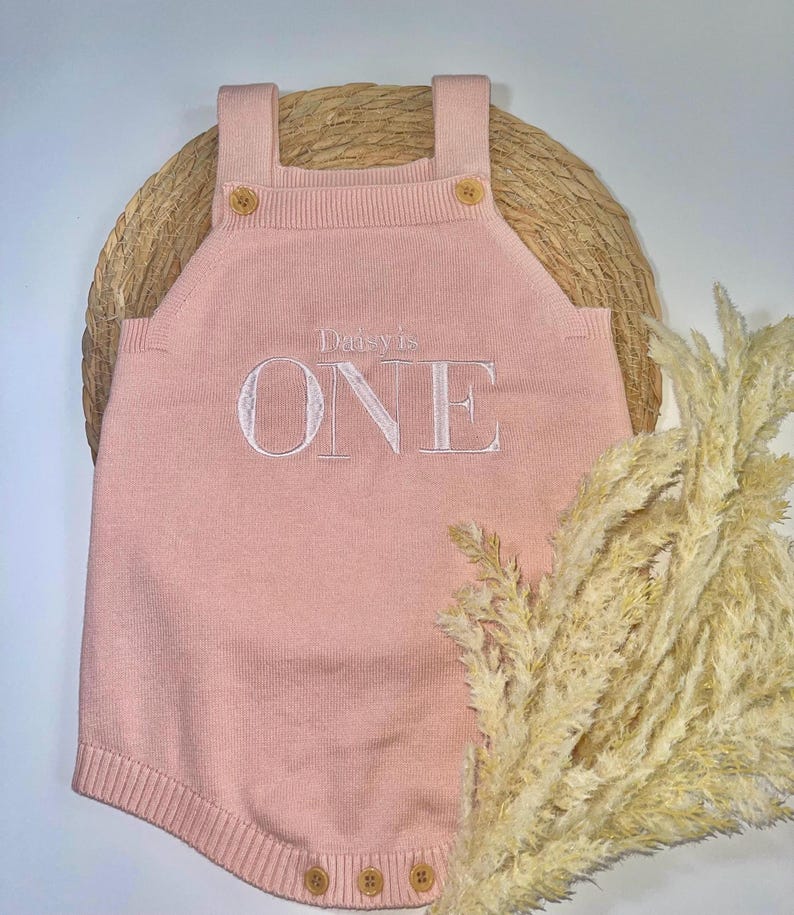 May include: A light pink knitted romper with the embroidered text "Daisy's ONE". The romper has wooden buttons on the straps and at the bottom. The romper is displayed on a woven mat with dried ornamental grass.