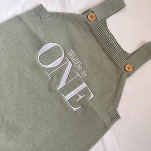 May include: Sage green knitted romper with the embroidered text "ONE" and "Wolfie is" in white. The romper features wooden buttons on the straps. A comfortable outfit for a baby's first birthday.