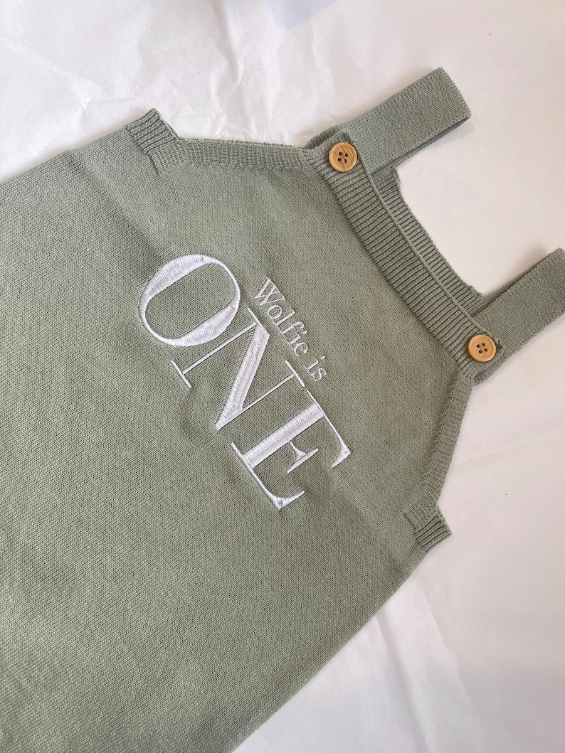 May include: Sage green knitted romper with the embroidered text "ONE" and "Wolfie is" in white. The romper features wooden buttons on the straps. A comfortable outfit for a baby's first birthday.