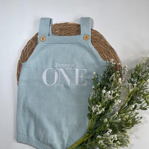 May include: A light blue knitted romper with the embroidered text "Lenny is ONE". The romper has wooden buttons on the straps and at the bottom. The romper is displayed on a woven mat with a sprig of white flowers.