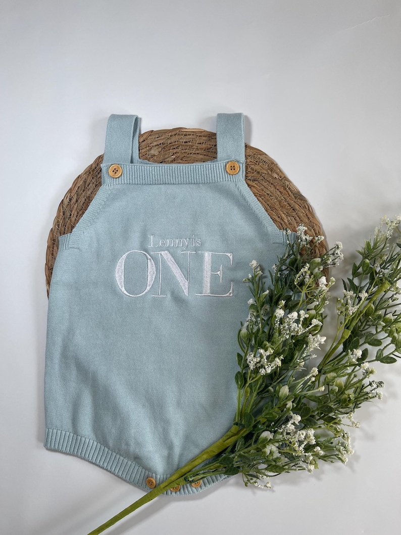 May include: A light blue knitted romper with the embroidered text "Lenny is ONE". The romper has wooden buttons on the straps and at the bottom. The romper is displayed on a woven mat with a sprig of white flowers.