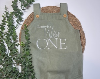 May include: Sage green baby romper with the embroidered text "Lenny is a Wild ONE." The romper features wooden buttons on the straps and along the bottom. The garment is made of a soft knit fabric and is designed for a baby.