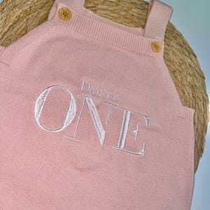 May include: A light pink knitted romper with shoulder straps and wooden buttons. The word "ONE" is embroidered in white, with "Daisy's is" above it. The romper is designed for a baby's first birthday.