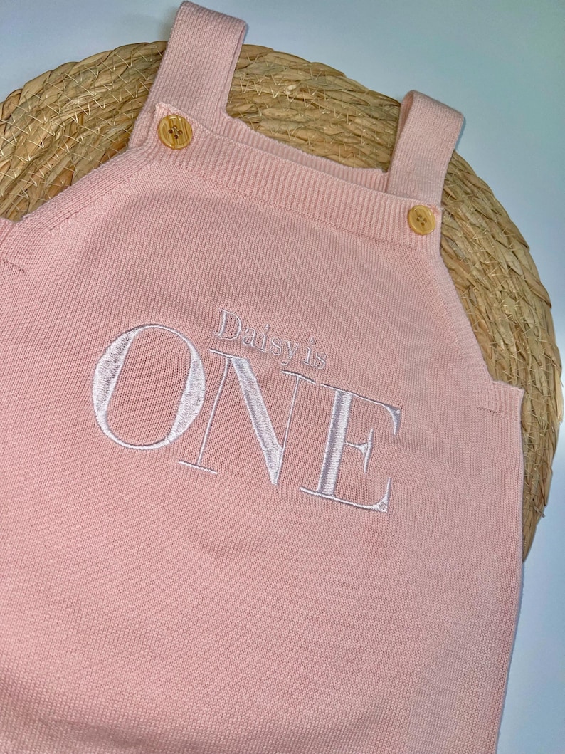 May include: A light pink knitted romper with shoulder straps and wooden buttons. The word "ONE" is embroidered in white, with "Daisy's is" above it. The romper is designed for a baby's first birthday.