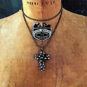Puede incluir: Un collar de cadena de plata con un colgante de cruz de plata. La cruz está adornada con pequeñas cuentas redondas. El collar está colgado de un maniquí. La etiqueta del collar dice "J.R. Designer-Artist-Sculptor Model Forming Model Forms Fur Designed & Factories 115 West 25th St. NY."