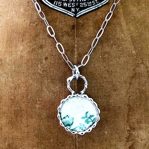 May include: A silver-toned necklace with a circular pendant. The pendant features a white background with a green floral design and a scalloped edge. The necklace has a chain link design.