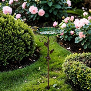 Puede incluir: Un bebedero para p&aacute;jaros de vidrio verde con un soporte de metal color bronce. El bebedero tiene un borde festoneado y est&aacute; lleno de agua. Un peque&ntilde;o p&aacute;jaro bebe del borde. El bebedero est&aacute; rodeado de arbustos verdes y rosas rosas.
