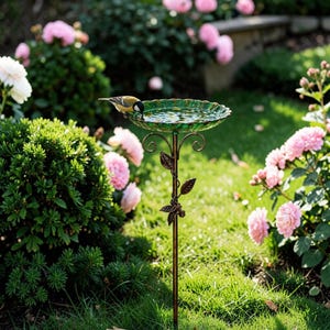 Peut inclure: Un abreuvoir décoratif avec une vasque verte et festonnée remplie d'eau. Il est soutenu par un pied en métal de couleur bronze avec des détails de feuilles et de vigne. Un petit oiseau boit au bord. Le tout est dans un jardin fleuri.