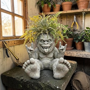 以下が含まれることがあります： 笑顔で舌を出した、ロックオンのハンドジェスチャーをした灰色のコンクリート製ビッグフットプランター。 プランターには緑と黄色の植物が植えられており、石の表面に置かれています。 植物愛好家のためのユニークな装飾アイテムです。
