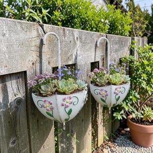 Op de afbeelding: Twee witte, parapluvormige hangende plantenbakken met roze en paarse bloemmotieven en vetplanten. De plantenbakken hangen aan een houten hek en tonen een charmante tuinesthetiek. De planten zijn een mix van groene vetplanten en kleurrijke bloemen.