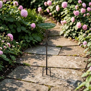 Puede incluir: Un bebedero decorativo para p&aacute;jaros con un cuenco de vidrio verde y un soporte de metal oscuro. El soporte presenta detalles de hojas y espirales. El bebedero est&aacute; situado en un camino de piedra rodeado de flores rosas y vegetaci&oacute;n.