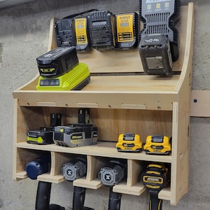 May include: A wooden power tool organizer mounted on a wall, holding various power tool batteries, chargers, and drills. The batteries are black and yellow, and the drills are black and gray. The organizer is made of light-colored wood.