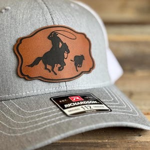 May include: A gray and white baseball cap with a brown leather patch featuring a silhouette of a cowboy roping a calf. The patch is stitched onto the front of the cap. The cap has a white mesh back and a black and white label that reads "EST. 1870 RICHARDSON 112 ORIGINAL TRUCKER YOUTH".