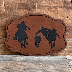 May include: A brown leather patch with a silhouette of three cowboys roping a calf. The cowboys are wearing hats and riding horses. The patch is perfect for adding a touch of Western flair to your projects.