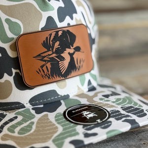 May include: A camouflage baseball cap featuring a rectangular tan leather patch on the front. The patch displays a black engraved illustration of a dog holding a duck in its mouth. A circular black sticker with the text BLACKHAWK is on the brim.