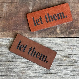 May include: Two rectangular brown leather patches with rounded corners, each featuring the text "let them." embossed in a bold, black, serif font. The patches have a smooth, consistent texture and are positioned diagonally against each other.