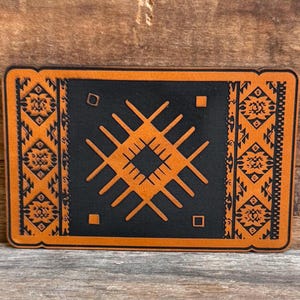 May include: A brown leather coaster with a black geometric design in the center and a brown patterned border.