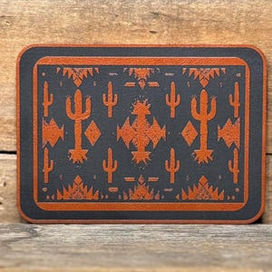 May include: A brown leather coaster with a black background and an orange cactus pattern. The pattern includes geometric shapes and cacti.