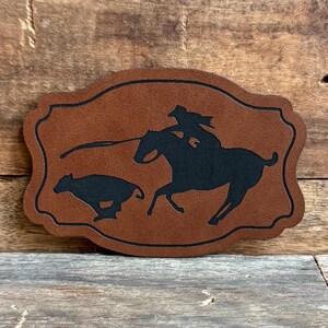 May include: A brown leather patch with a silhouette of a cowgirl riding a horse and roping a calf.