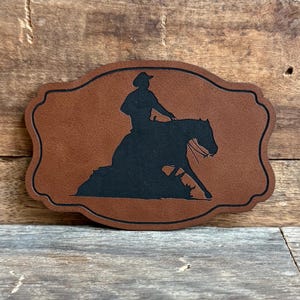 May include: A brown leather patch with a black silhouette of a rider and horse in a reining pose.