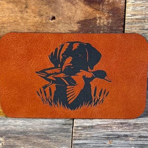 May include: A rectangular, orange leather patch featuring a black silhouette of a Labrador Retriever holding a duck in its mouth. The dog is positioned above a depiction of reeds.
