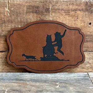 May include: A brown leather patch with a black silhouette of a cowboy riding a horse and a calf running in front of them.