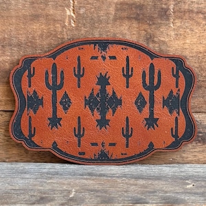 May include: A brown leather belt buckle with a desert cactus design. The buckle has a rectangular shape with rounded edges and a black border. The cactus and geometric patterns are in black.