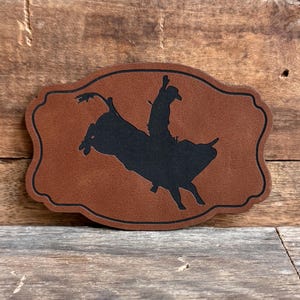 May include: A brown leather patch with a black silhouette of a rodeo rider on a bucking bull. The patch has a decorative border and is set against a wooden background.