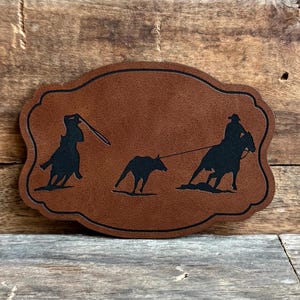 May include: A brown leather patch with a silhouette of two cowboys on horseback roping a calf. The patch has a decorative scalloped edge.