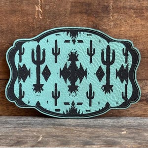 May include: A turquoise-colored, leather-like belt buckle with a black southwestern design. The design features cacti, diamonds, and geometric shapes. The buckle has a decorative, scalloped edge and is set against a wooden background.