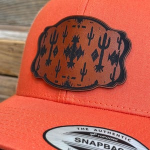 May include: An orange baseball cap with a leather patch featuring a desert cactus design. The patch is brown with black cactus and geometric shapes. The hat has a curved brim and a snapback closure. The label reads "SNAPBACK".