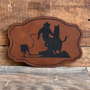 May include: A brown leather patch with a black silhouette of a cowboy roping a calf. The patch is shaped like a traditional Western saloon sign.