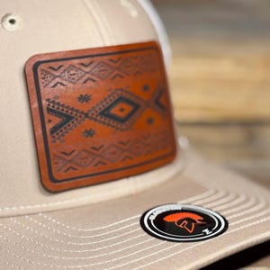 May include: A beige baseball cap featuring a rectangular brown leather patch on the front panel. The patch is embossed with a dark brown geometric pattern. A circular black sticker with an orange helmet icon and text is on the brim.