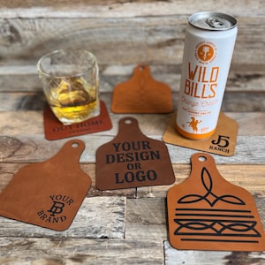 May include: A collection of tan leather coasters shaped like livestock ear tags, featuring various black laser-engraved designs including text and logos. One coaster supports a glass of amber liquid, while another holds a tall white soda can.