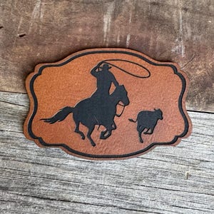 May include: A brown leather patch with a silhouette of a cowboy riding a horse and roping a calf. The patch is shaped like a rounded rectangle with a decorative border.