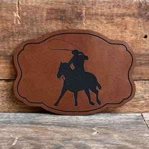 May include: A brown leather patch with a black silhouette of a cowboy riding a horse and lassoing. The patch has a decorative scalloped edge.