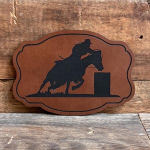 May include: A brown leather patch with a black silhouette of a person riding a horse jumping over a barrel. The patch is shaped like a rounded rectangle with a decorative border.