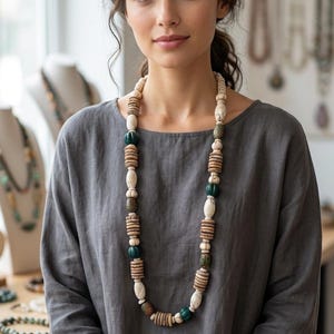 Könnte beinhalten: Eine lange Perlenkette mit verschiedenen Naturtönen, darunter Creme, Braun und Grün. Die Kette wird von einer Person mit einem grauen Oberteil getragen. Weitere Halsketten sind im Hintergrund zu sehen.