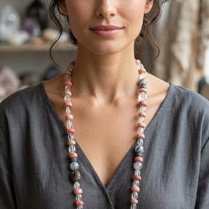 Può includere: Una lunga collana di perline con pietre e perline rosa, trasparenti, grigie e nere. La collana è indossata da una persona che indossa un top grigio.
